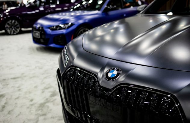 Frente de um BMW cinza fosco com logo em destaque. Dois carros desfocados, azul e roxo, estão ao fundo.