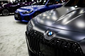 Frente de um BMW cinza fosco com logo em destaque. Dois carros desfocados, azul e roxo, estão ao fundo.