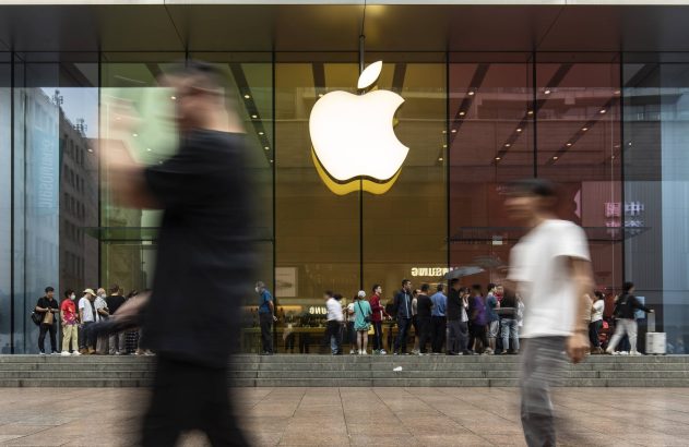 Loja Apple com logo luminoso em fachada de vidro. Pessoas em desfoque passam e outras esperam em fila nas escadas.