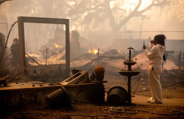 Mulher fotografa cenário de destruição pós-incêndio. Fumaça, chamas e escombros de casas ao redor.