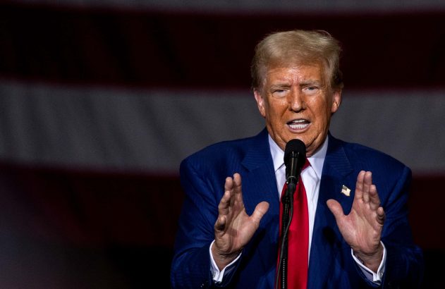 Donald Trump discursa com microfone e mãos gesticulando, vestido com terno azul e gravata vermelha, sobre fundo de bandeira.