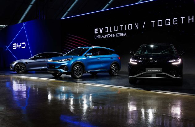 Três carros BYD (azul, preto, cinza) em exibição em seu lançamento na Coreia. Painel 'BYD LAUNCH IN KOREA'.
