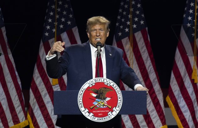 Donald Trump discursa em pódio da NRA, gesticulando, com bandeiras americanas ao fundo.