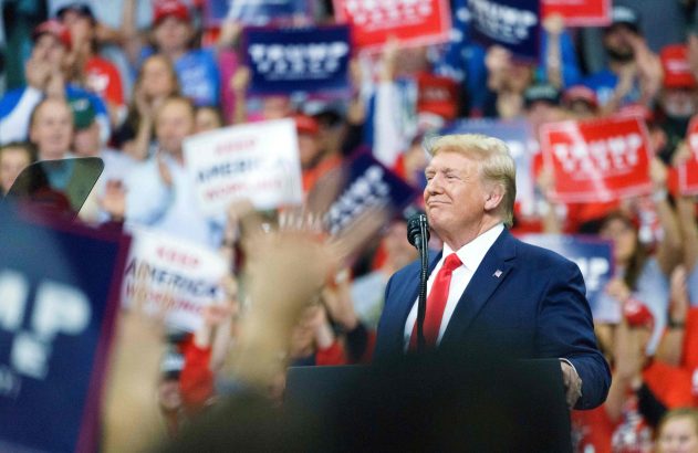Donald Trump sorri no pódio em um comício, com multidão vibrante segurando cartazes de apoio.