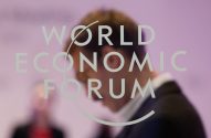 Texto "WORLD ECONOMIC FORUM" em primeiro plano. Ao fundo, um homem desfocado de terno olhando para baixo.