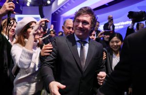 Javier Milei, de terno, sorri cercado por pessoas filmando e fotografando em reunião.