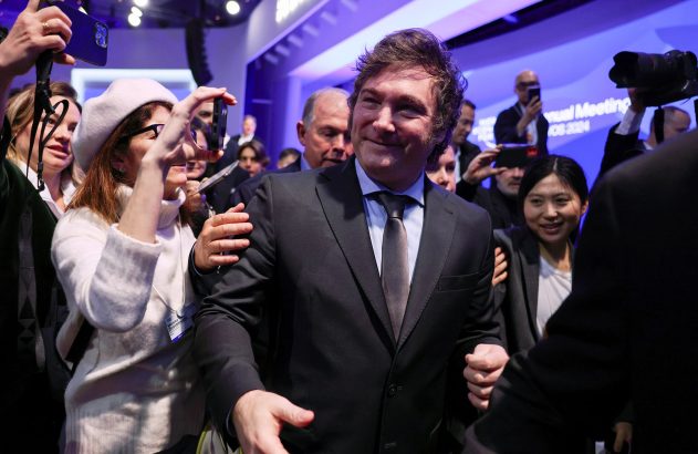 Javier Milei, de terno, sorri cercado por pessoas filmando e fotografando em reunião.