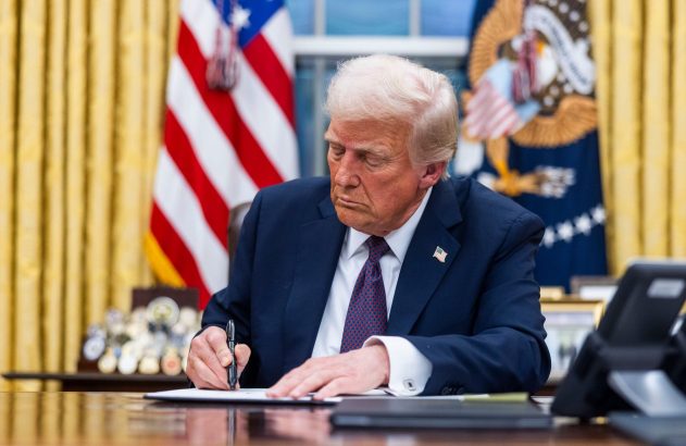 Donald Trump assina documento em mesa. Bandeira dos EUA e o selo presidencial ao fundo.