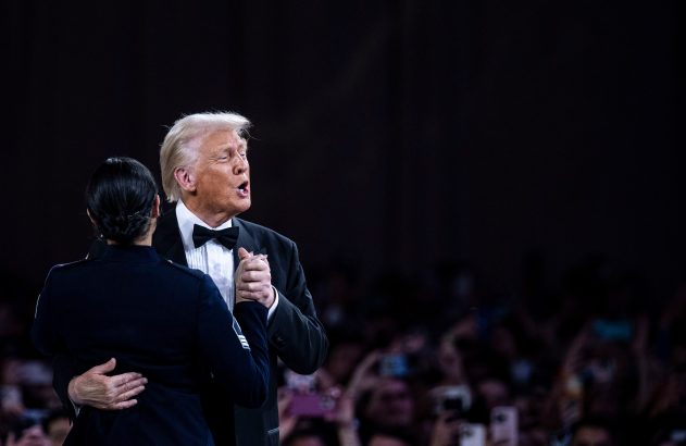 Donald Trump de smoking e mulher em uniforme militar se abraçam dançando. Ele está com a boca aberta. Pessoas com celulares ao fundo.