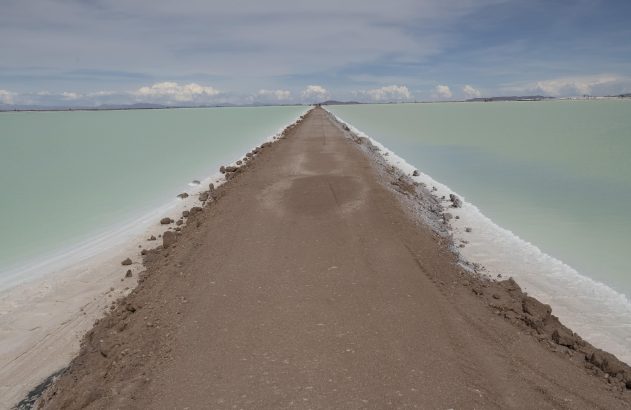 Estrada de terra reta dividindo lago salgado verde-claro, com bordas de sal. Montanhas e céu nublado ao fundo.