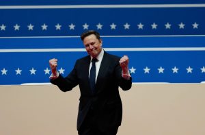 Elon Musk, de terno, comemora com punhos erguidos à frente de um fundo azul com listras e estrelas brancas.