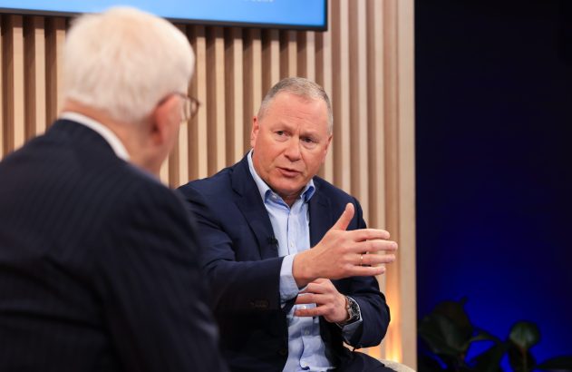 Em um estúdio, um homem de terno azul gesticula e fala para outro homem, visto de costas.