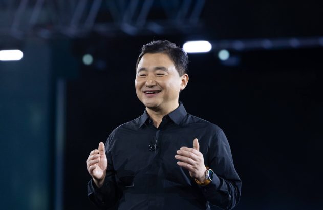Homem asiático sorrindo e gesticulando em palco escuro, usando camisa preta e relógio.