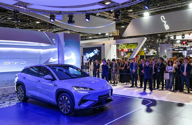 BYD Sealion 7 roxo em estande de feira, iluminado em azul. Pessoas observam o carro em exibição.