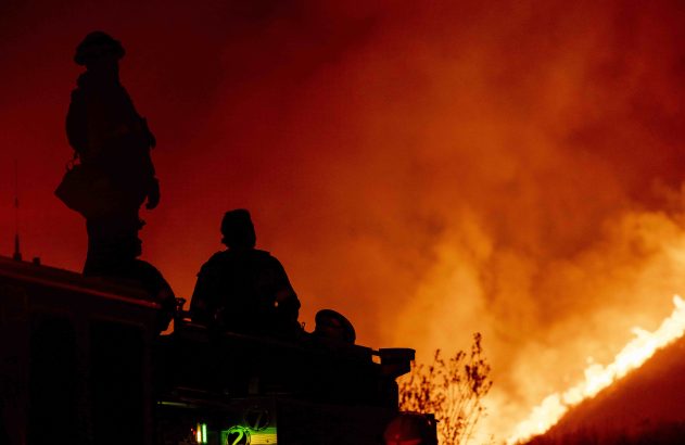 Silhuetas de bombeiros sobre caminhão em meio a um incêndio florestal, com o céu iluminado de vermelho e laranja.