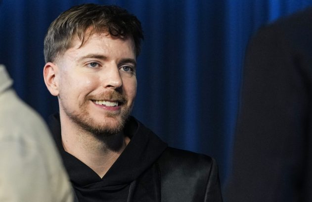 Homem de barba e cabelo castanho sorrindo e olhando para o lado. Veste preto, em fundo azul escuro desfocado.