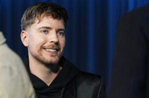 Homem de barba e cabelo castanho sorrindo e olhando para o lado. Veste preto, em fundo azul escuro desfocado.