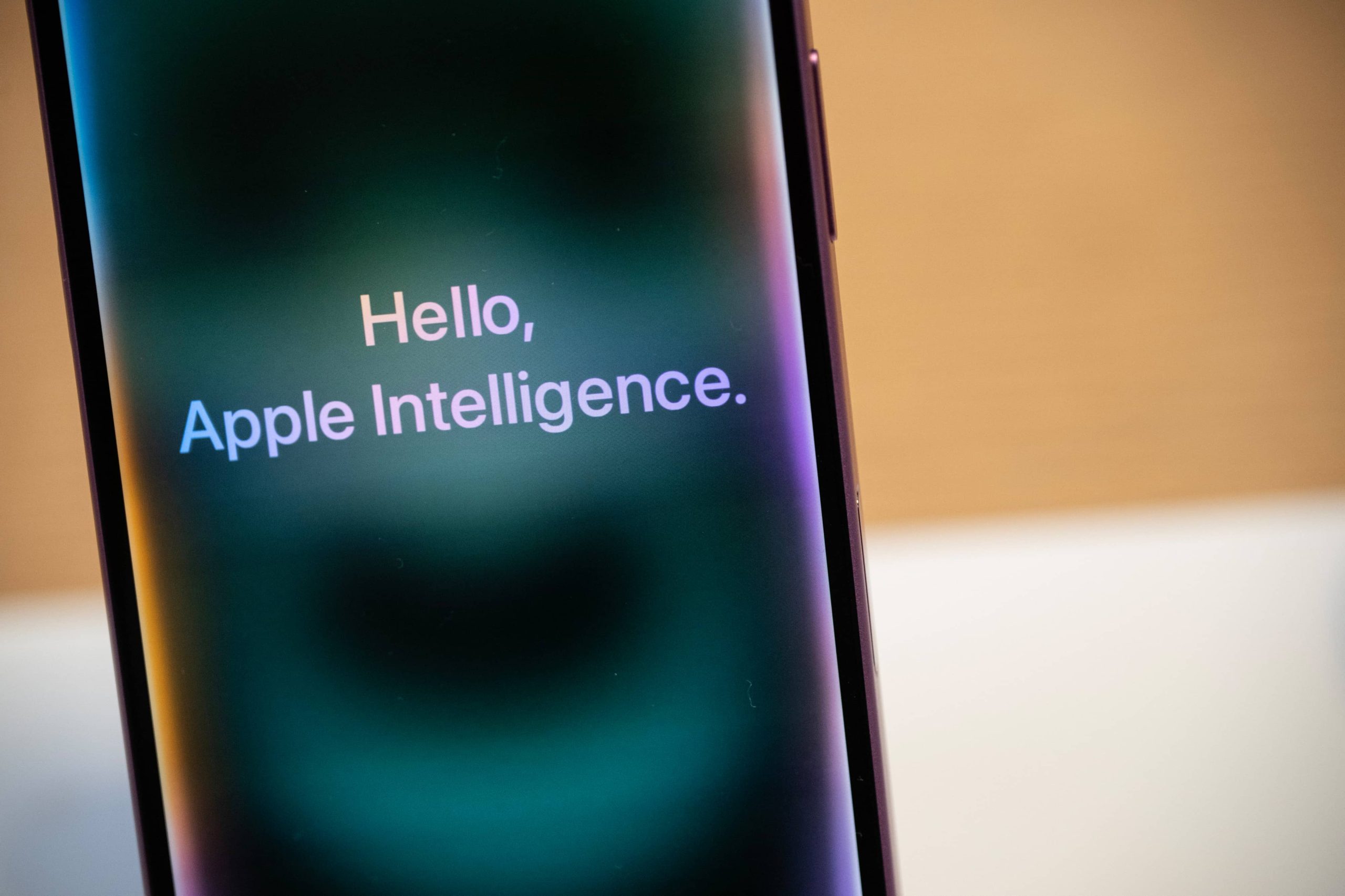 Celular com texto 'Hello, Apple Intelligence' na tela, sobre fundo turvo.