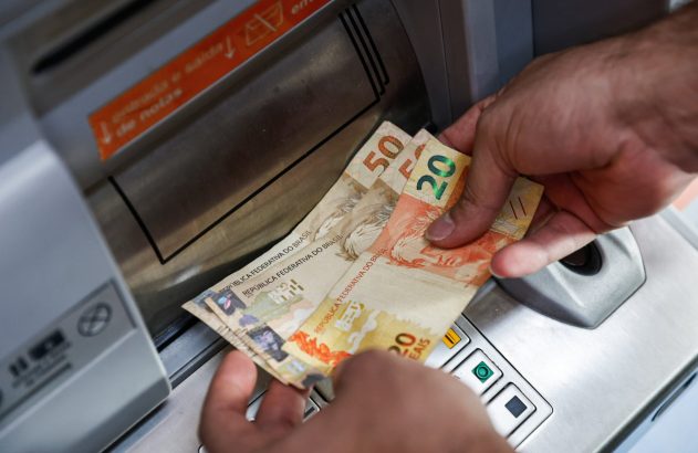 Mãos retiram cédulas de 20 e 50 Reais de um caixa eletrônico no Brasil.
