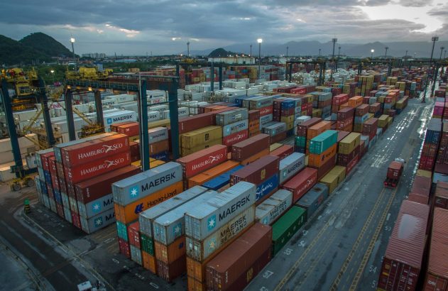 Vista aérea de um porto com pilhas de contêineres coloridos, guindastes e um caminhão sob céu nublado.