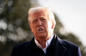 Donald Trump, com a boca aberta, discursa. Veste casaco escuro e camisa branca. Fundo externo desfocado.