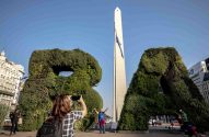 Mulher fotografa outra posando com topiarias 'BA' e Obelisco de Buenos Aires ao fundo, sob céu azul.