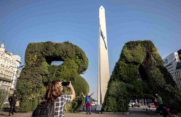 Mulher fotografa outra posando com topiarias 'BA' e Obelisco de Buenos Aires ao fundo, sob céu azul.