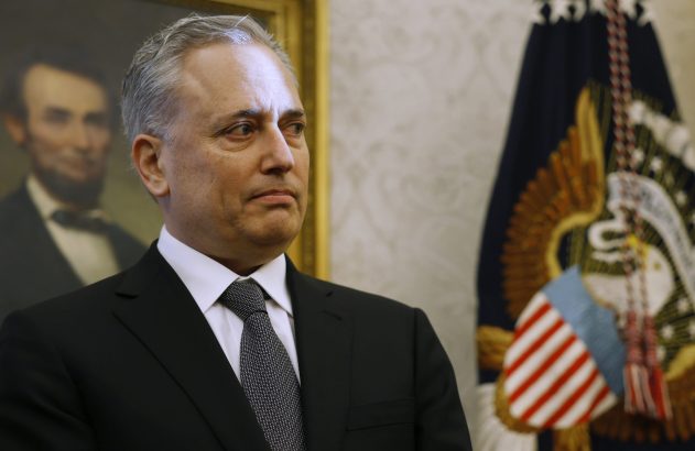 Homem de terno preto e cabelo grisalho olha. Ao fundo, retrato de Lincoln e selo presidencial dos EUA.