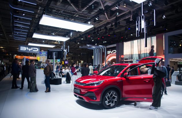 Carro BYD vermelho em exposição. Portas abertas, pessoas interagem.