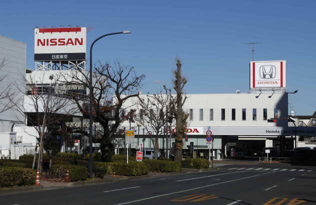 Fachadas das concessionárias Nissan e Honda, com rua, árvores e uma pessoa andando na calçada em um dia claro.