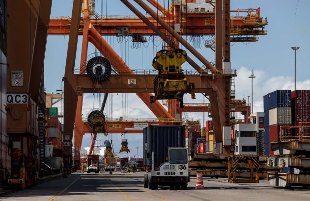 Terminal de contêineres movimentado: guindastes laranjas içam contêineres e um caminhão carrega um.