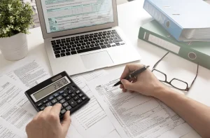 Mãos preenchendo formulário 1120, usando calculadora. Laptop com formulário fiscal, óculos e pastas em mesa.