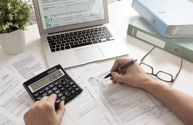 Mãos preenchendo formulário 1120, usando calculadora. Laptop com formulário fiscal, óculos e pastas em mesa.