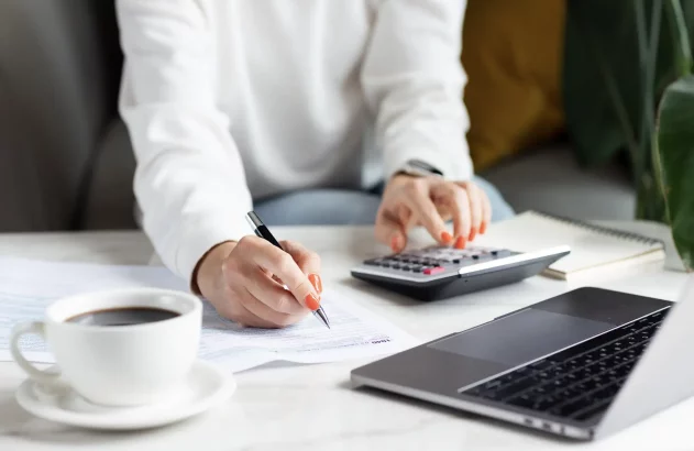 Pessoa preenche declaração de imposto de renda, usando calculadora e caneta. Na mesa, laptop e xícara de café.