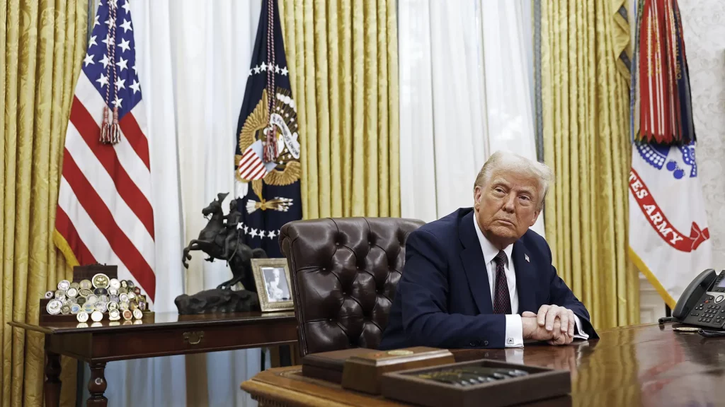 O presidente dos EUA, Donald Trump, em terno escuro, está sentado no Salão Oval, com bandeiras dos EUA e do Exército ao fundo, adornado com medalhas e uma estátua de cavalo na mesa.