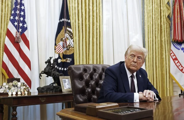 O presidente dos EUA, Donald Trump, em terno escuro, está sentado no Salão Oval, com bandeiras dos EUA e do Exército ao fundo, adornado com medalhas e uma estátua de cavalo na mesa.