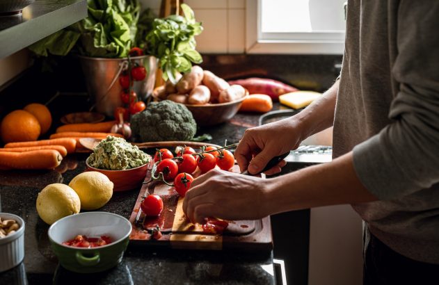 Pessoa fatiando tomate cereja em tábua de corte. Bancada de cozinha com diversos legumes frescos para preparar uma refeição.