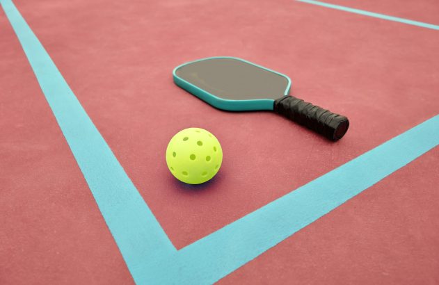 Uma raquete de pickleball e uma bola amarela perfurada em uma quadra vermelha com linhas azuis.