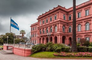 A Casa Rosada, edifício rosa, com bandeira da Argentina e jardim sob céu nublado.