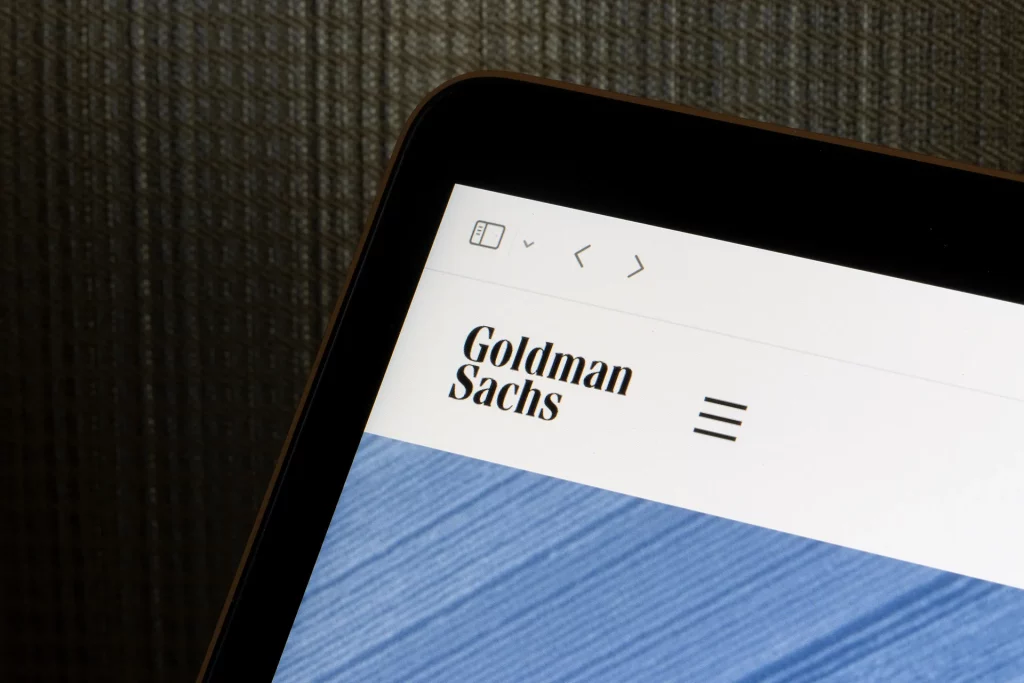 A imagem mostra a tela de um dispositivo eletrônico exibindo o nome "Goldman Sachs" em letras pretas com um menu de navegação ao lado, sobre um fundo branco e uma faixa azul texturizada.