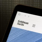 A imagem mostra a tela de um dispositivo eletrônico exibindo o nome "Goldman Sachs" em letras pretas com um menu de navegação ao lado, sobre um fundo branco e uma faixa azul texturizada.