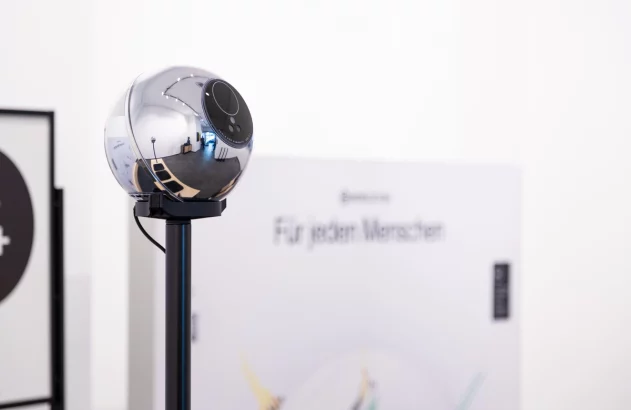 Câmera esférica cromada em suporte preto, refletindo o ambiente. Fundo desfocado com texto "Für jeden Menschen".