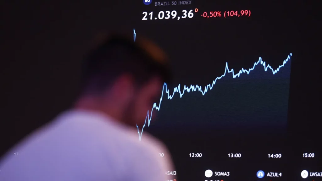 Gráfico de linhas mostrando a queda de 0,50% no índice Brazil 50, com uma figura humana desfocada em frente à tela exibindo variações de preços e logotipos de empresas na parte inferior.