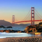 A paisagem do entardecer na Ponte Golden Gate destaca-se com céu em tons de azul e laranja, mar sereno, praia com rochas e vegetação dourada ao fundo.