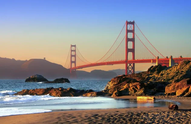 A paisagem do entardecer na Ponte Golden Gate destaca-se com céu em tons de azul e laranja, mar sereno, praia com rochas e vegetação dourada ao fundo.