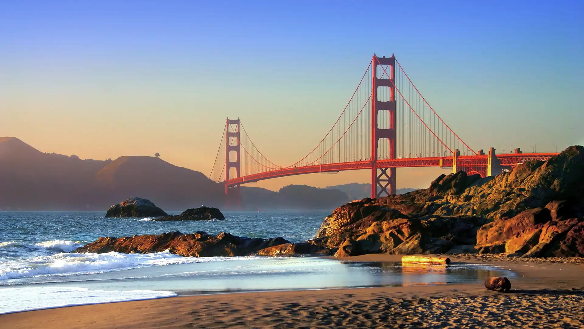 A paisagem do entardecer na Ponte Golden Gate destaca-se com céu em tons de azul e laranja, mar sereno, praia com rochas e vegetação dourada ao fundo.