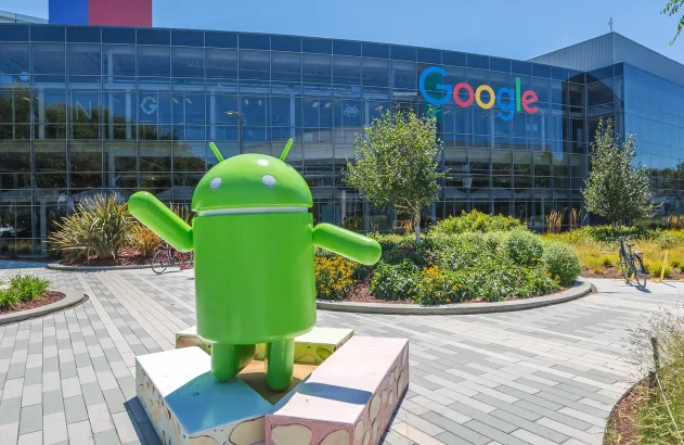 Prédio moderno de vidro com logomarca do Google, estátua verde do Android, jardins bem cuidados e bicicleta estacionada, em um dia ensolarado.