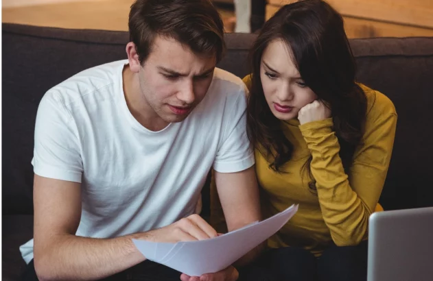 Homem e mulher com expressões sérias analisam documentos. Há um laptop na mesa, sugerindo gerenciamento de finanças.