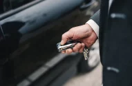 Uma mão segurando uma chave moderna em frente a um carro preto, sugerindo ação de destrancar ou trancar o veículo.