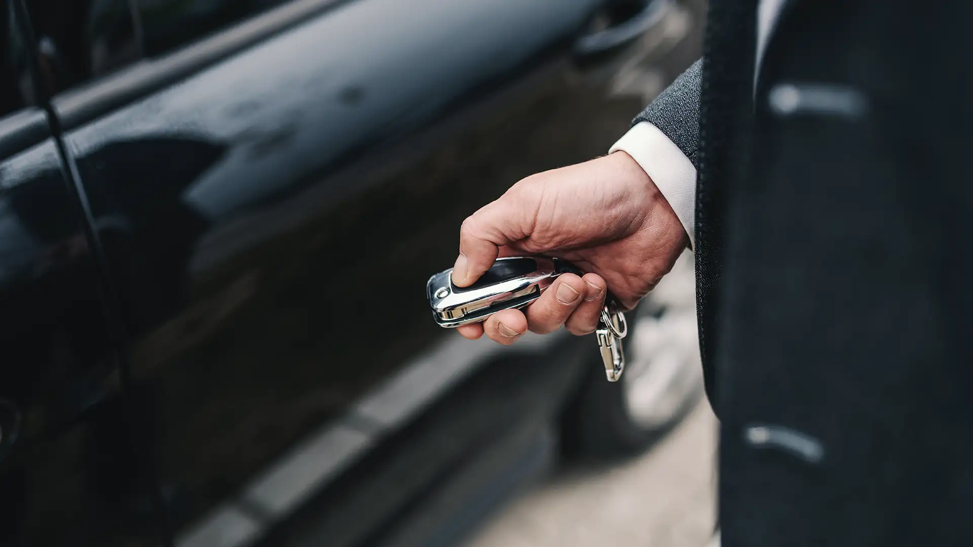 Uma mão segurando uma chave moderna em frente a um carro preto, sugerindo ação de destrancar ou trancar o veículo.